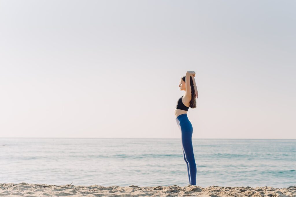 woman in black brassier and blue leggings standing on seashore doing yoga